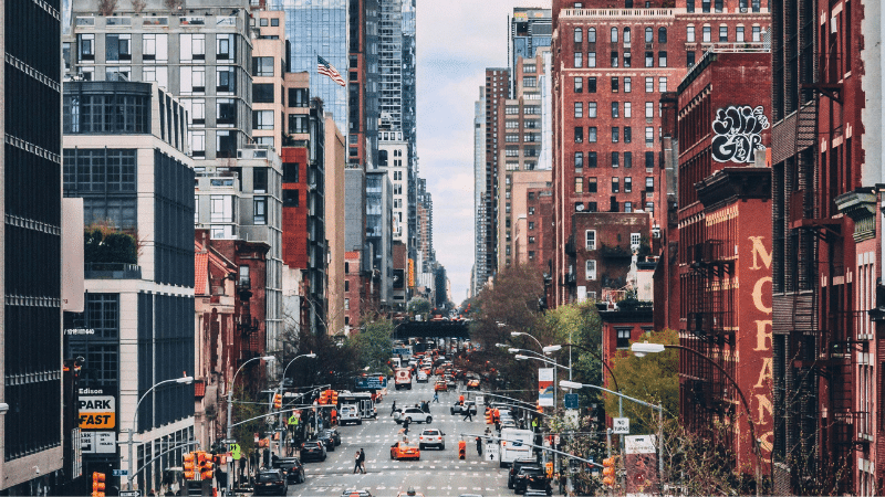 A bustling city street, reminiscent of Digilogue Days, is lined with tall buildings. Numerous cars and pedestrians navigate the urban landscape, while a cloudy sky looms above, capturing the harmonious blend of digital hustle and timeless urban rhythm.