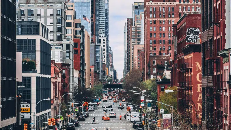 A bustling city street, reminiscent of Digilogue Days, is lined with tall buildings. Numerous cars and pedestrians navigate the urban landscape, while a cloudy sky looms above, capturing the harmonious blend of digital hustle and timeless urban rhythm.