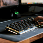 A person types on a mechanical keyboard with backlit keys, set on a large mouse pad, in front of two computer monitors.