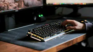 A person types on a mechanical keyboard with backlit keys, set on a large mouse pad, in front of two computer monitors.