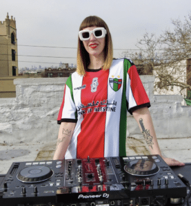 A person wearing a Palestine-themed shirt and sunglasses stands behind a DJ controller on a rooftop.