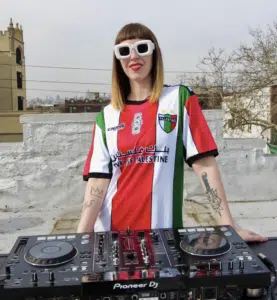 A person wearing a Palestine-themed shirt and sunglasses stands behind a DJ controller on a rooftop.