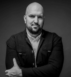 A bald man with a beard, exuding the confident aura of an A&R executive, poses with arms crossed in a dark jacket and light shirt against a shadowy backdrop.