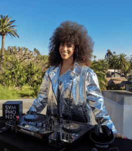 A person with curly hair wearing a silver jacket stands outdoors behind DJ equipment with palm trees in the background.