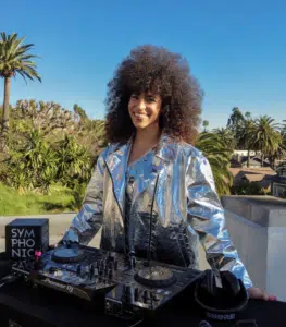 A person with curly hair wearing a silver jacket stands outdoors behind DJ equipment with palm trees in the background.