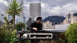 A DJ wearing headphones performs on a rooftop, where your story starts, surrounded by plants and urban buildings against a backdrop of cloudy skies.