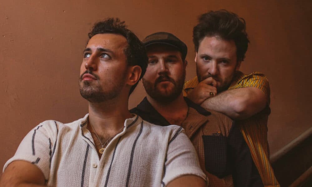 Three men in casual attire stand together on a staircase, looking in different directions with thoughtful expressions, as if pondering the inspiration behind their new music.