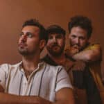 Three men in casual attire stand together on a staircase, looking in different directions with thoughtful expressions, as if pondering the inspiration behind their new music.