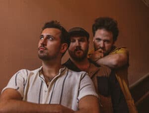 Three men in casual attire stand together on a staircase, looking in different directions with thoughtful expressions, as if pondering the inspiration behind their new music.