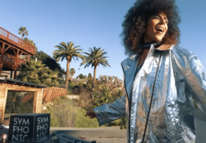 Person in a shiny silver jacket stands and smiles outdoors near palm trees and wooden structures on a sunny day.