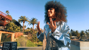 A person in a shiny silver jacket smiles brightly, as if part of a cherished story, while standing outdoors with palm trees swaying gently in the background.