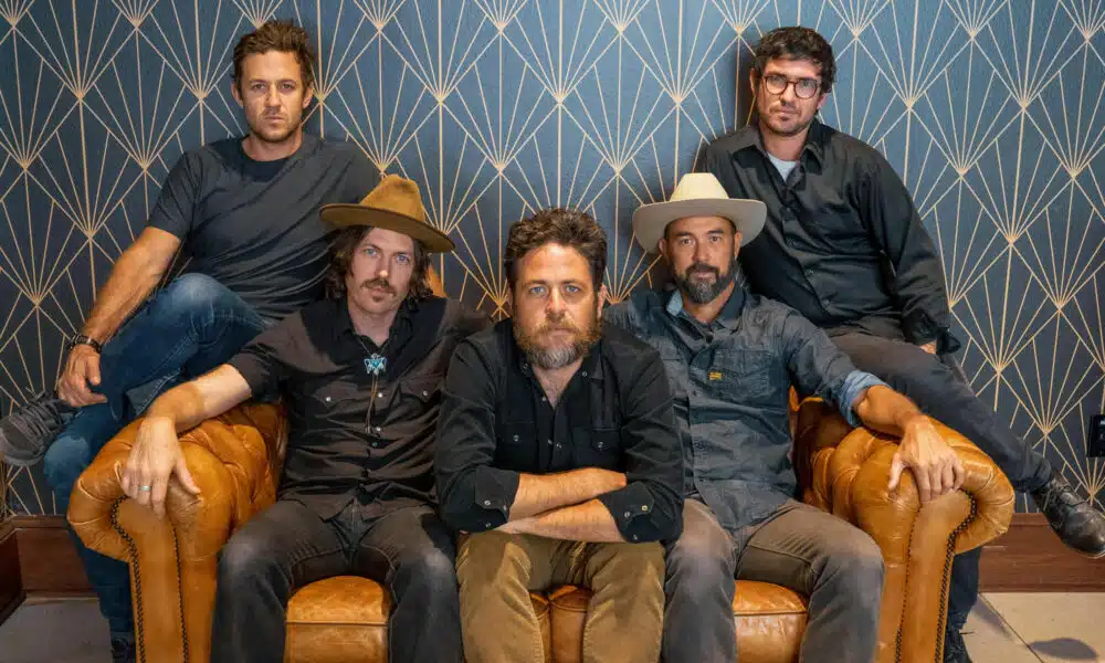 Five men sit and stand around a leather couch, posing for a group photo. The background features a geometric patterned wall, setting the stage for their new music release.