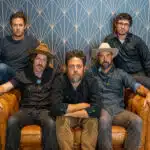 Five men sit and stand around a leather couch, posing for a group photo. The background features a geometric patterned wall, setting the stage for their new music release.