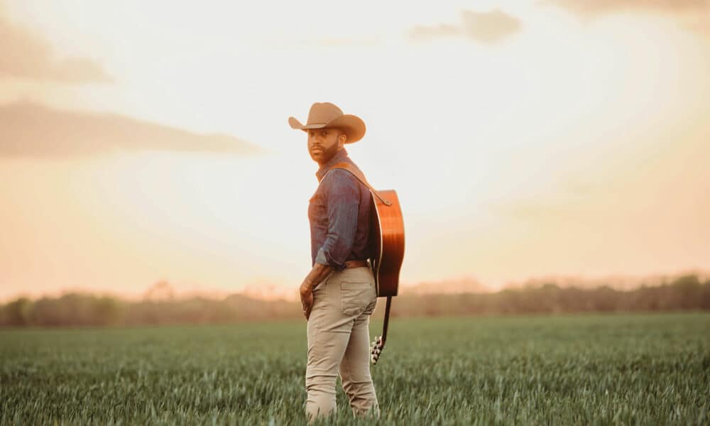 A man in a cowboy hat stands in a field with a guitar on his back, facing the camera. As the clear sky meets the grassy landscape at sunset, he seems ready to share new music inspired by this serene setting.