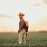 A man in a cowboy hat stands in a field with a guitar on his back, facing the camera. As the clear sky meets the grassy landscape at sunset, he seems ready to share new music inspired by this serene setting.