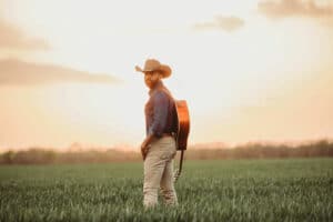 A man in a cowboy hat stands in a field with a guitar on his back, facing the camera. As the clear sky meets the grassy landscape at sunset, he seems ready to share new music inspired by this serene setting.