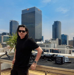 Person in black shirt stands on a rooftop with DJ equipment, overlooking a cityscape with tall buildings under a blue sky.