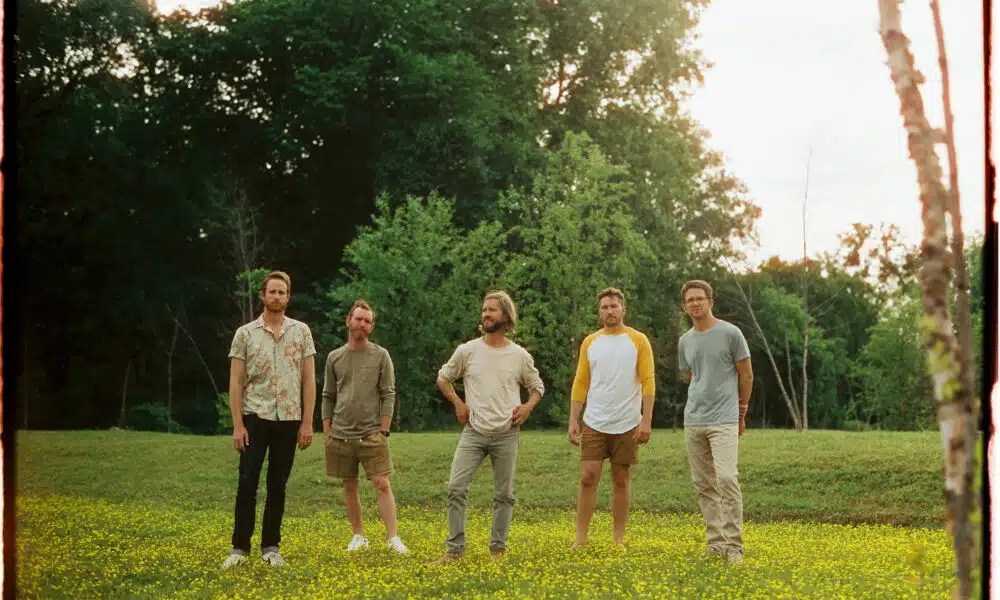 Five men stand in a field of yellow flowers with trees in the background, ready to share their new music with the world.