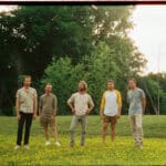 Five men stand in a field of yellow flowers with trees in the background, ready to share their new music with the world.