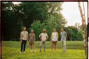 Five men stand in a field of yellow flowers with trees in the background, ready to share their new music with the world.