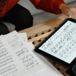 A person reviews sheet music—perhaps holiday songs you can cover for free—on paper and a tablet, with a pen in hand, at a wooden table.