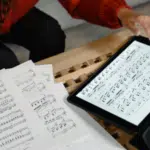 A person reviews sheet music—perhaps holiday songs you can cover for free—on paper and a tablet, with a pen in hand, at a wooden table.