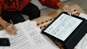 A person reviews sheet music—perhaps holiday songs you can cover for free—on paper and a tablet, with a pen in hand, at a wooden table.