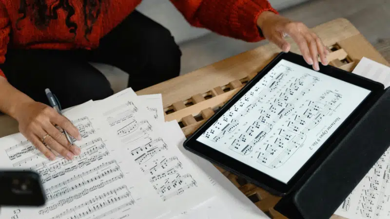 A person reviews sheet music—perhaps holiday songs you can cover for free—on paper and a tablet, with a pen in hand, at a wooden table.