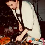 A DJ in a light-colored jacket operates turntables and a mixer at an indoor event, surrounded by vinyl records, party decorations, and hints of marketing on Instagram displayed near the setup.
