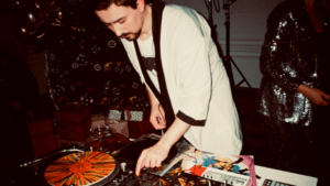 A DJ in a light-colored jacket operates turntables and a mixer at an indoor event, surrounded by vinyl records, party decorations, and hints of marketing on Instagram displayed near the setup.
