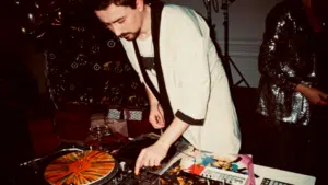 A DJ in a light-colored jacket operates turntables and a mixer at an indoor event, surrounded by vinyl records, party decorations, and hints of marketing on Instagram displayed near the setup.