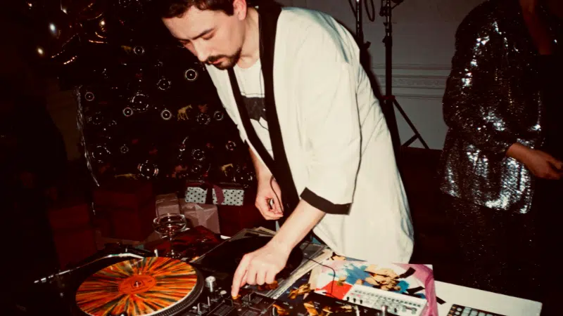 A DJ in a light-colored jacket operates turntables and a mixer at an indoor event, surrounded by vinyl records, party decorations, and hints of marketing on Instagram displayed near the setup.