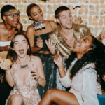 A group of people dressed in party attire laugh and celebrate the holidays together indoors, holding props and drinks, with a retro patterned wall in the background.