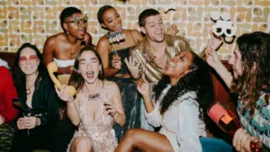 A group of people dressed in party attire laugh and celebrate the holidays together indoors, holding props and drinks, with a retro patterned wall in the background.