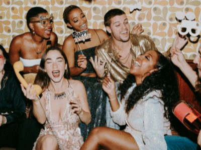 A group of people dressed in party attire laugh and celebrate the holidays together indoors, holding props and drinks, with a retro patterned wall in the background.