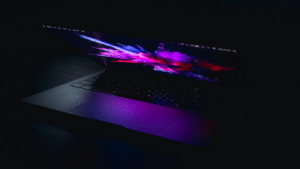 Introducing New SplitShare Updates in the SMS A partially open laptop with a colorful, abstract display screen, emitting a purple glow in a dark environment, captures the essence of innovation. The scene resembles the dynamic visual experience that SplitShare aims to deliver through seamless content sharing.