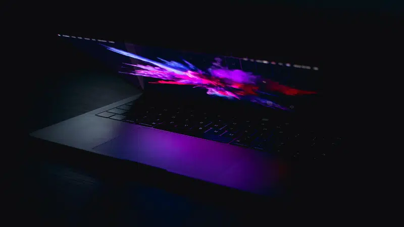 A partially open laptop with a colorful, abstract display screen, emitting a purple glow in a dark environment, captures the essence of innovation. The scene resembles the dynamic visual experience that SplitShare aims to deliver through seamless content sharing.