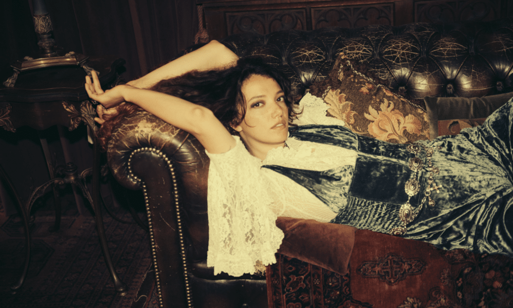 A person in vintage-style clothing reclines on a leather couch, surrounded by decorative pillows, with their arms resting behind their head, while new music plays softly in the background.
