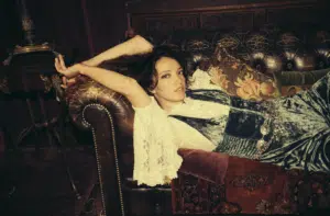 A person in vintage-style clothing reclines on a leather couch, surrounded by decorative pillows, with their arms resting behind their head, while new music plays softly in the background.