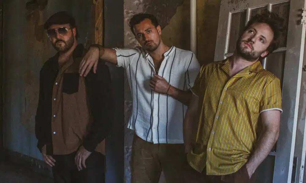 Three men stand casually against a rustic wall; one leans on a door, another rests his hand on a friend's shoulder, and the third, excited about new music, has hands in pockets.