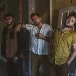 Three men stand casually against a rustic wall; one leans on a door, another rests his hand on a friend's shoulder, and the third, excited about new music, has hands in pockets.