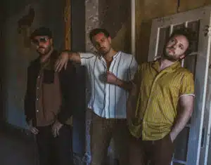 Three men stand casually against a rustic wall; one leans on a door, another rests his hand on a friend's shoulder, and the third, excited about new music, has hands in pockets.