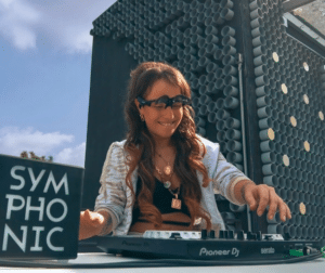 A person with long brown hair is DJ'ing outdoors, wearing sunglasses and headphones. A sign reads "Symphonic.