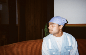 A person wearing a purple bandana and denim vest sits on an orange couch with closed eyes.