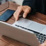 Close-up of a person's hands using a laptop on a wooden table, with a smartphone and a notebook nearby.