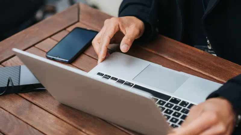 Close-up of a person's hands using a laptop on a wooden table, with a smartphone and a notebook nearby.