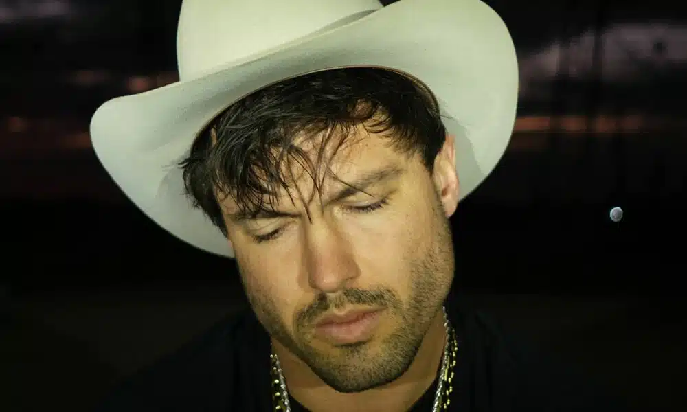 A man wearing a white cowboy hat and gold chain, with his eyes closed, stands in a dimly lit setting. The atmosphere buzzes with anticipation as he's lost in thought, perhaps dreaming of his new music debut.