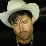 A man wearing a white cowboy hat and gold chain, with his eyes closed, stands in a dimly lit setting. The atmosphere buzzes with anticipation as he's lost in thought, perhaps dreaming of his new music debut.