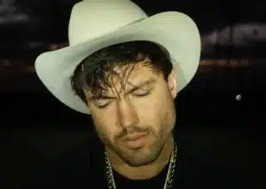A man wearing a white cowboy hat and gold chain, with his eyes closed, stands in a dimly lit setting. The atmosphere buzzes with anticipation as he's lost in thought, perhaps dreaming of his new music debut.