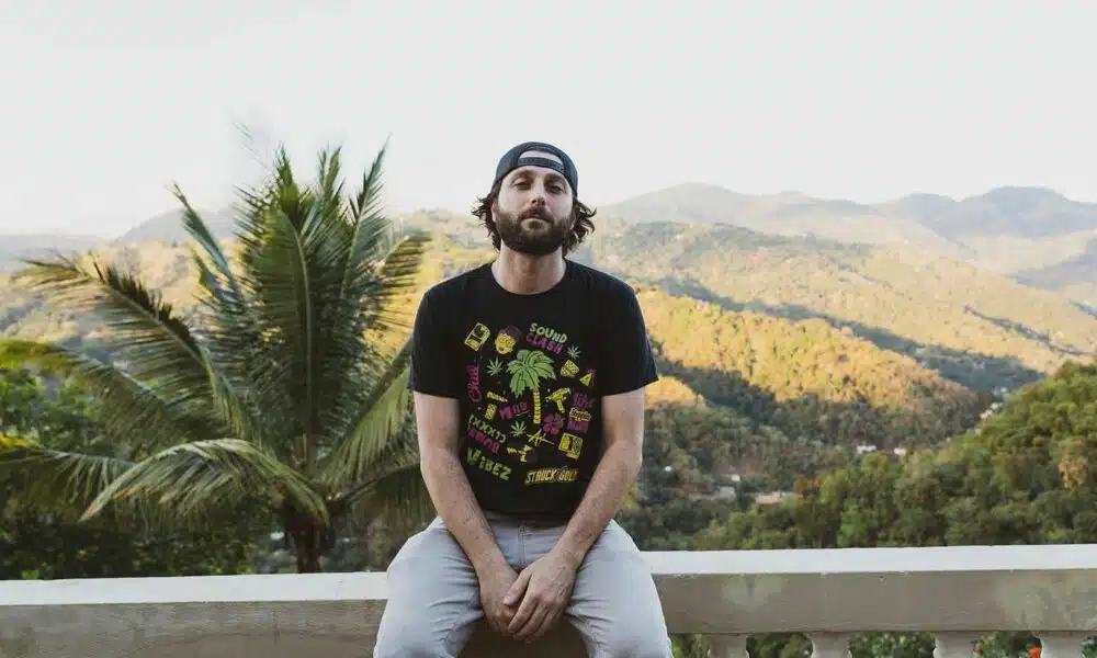 A person in a black t-shirt and cap sits on a ledge with a scenic mountain view in the background, creating the perfect spot for finding inspiration for new music.
